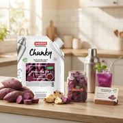Andros Chunky purple sweet potato sauce mix shown in a kitchen setting with a shaker bottle, a jar of sliced purple sweet potatoes, a prepared drink, and raw sweet potatoes on a wooden counter.